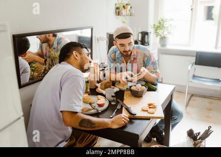 Coppia gay che condivide uno smartphone con l'altro mentre si mangia al tavolo da pranzo a casa Foto Stock