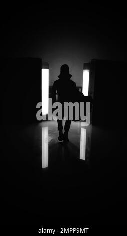 Scala di grigi verticale di una silhouette di una persona in un vestito che cammina verso un riflettore Foto Stock