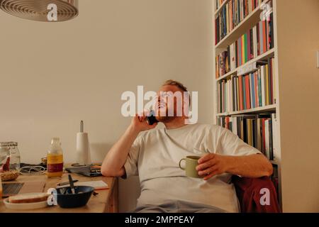 Uomo in sovrappeso che parla sullo smartphone tenendo la tazza mentre si siede a casa Foto Stock