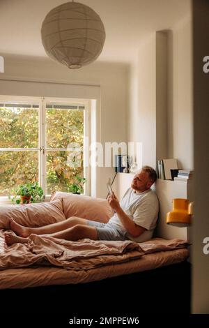 Uomo in sovrappeso che legge il libro mentre si siede sul letto a casa Foto Stock