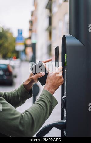 Mani di un uomo che effettua il pagamento tramite cellulare alla stazione di ricarica Foto Stock