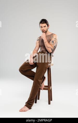 Un uomo elegante e tatuato che tocca il mento seduto su sfondo grigio Foto Stock