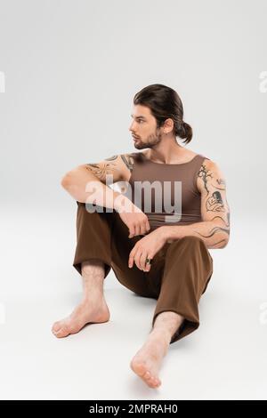 Vista laterale di un uomo dai capelli lunghi e tatuati seduto su sfondo grigio Foto Stock