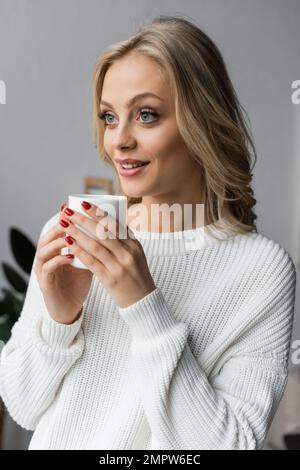 giovane donna bionda in maglione bianco che tiene una tazza di tè e guardando lontano a casa, immagine stock Foto Stock