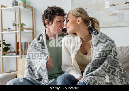 felice giovane uomo e donna bionda sorridente coperto di coperta seduta sul divano, immagine stock Foto Stock