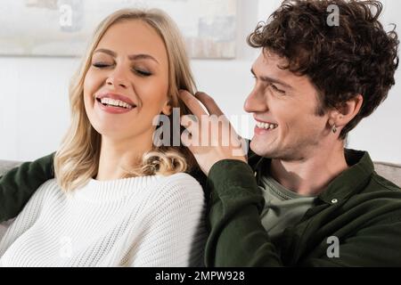 felice e riccio uomo in camicia regolazione biondo capelli di bella ragazza in maglione bianco, immagine stock Foto Stock