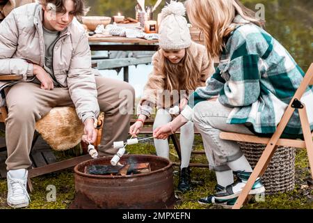 Bambini, famiglia, sorella fratello friggere marshmallows sul fuoco. Picnic nella natura. Sedia di legno, table.barbecue barbecue cibo. Viaggio di vacanza, fine settimana di vacanza dinne Foto Stock