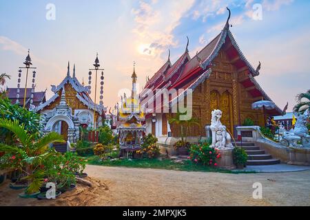 Il cielo nuvoloso tramonto sopra i santuari ornati del medievale Wat Mahawan, circondato da giardino fiorito, Chiang mai, Thailandia Foto Stock