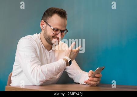 L'elegante giovane uomo d'affari europeo si siede a un tavolo in un bar e mostra il suo orologio intelligente attraverso lo schermo di uno smartphone. Foto Stock