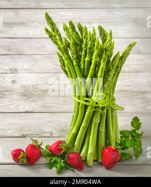 Mazzo di asparagi crudi verdi decorati con fragole fresche su sfondo di legno chiaro e luminoso, formato verticale Foto Stock