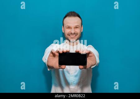 Elegante e positivo sorridente giovane simpatico uomo d'affari d'Europa in una camicia bianca tiene un telefono cellulare su sfondo blu. Foto Stock