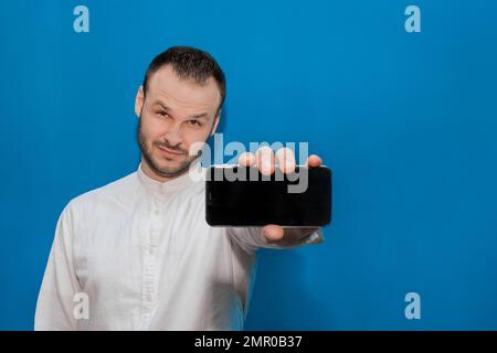 Elegante, giovane uomo d'affari caucasico aspetto in una camicia bianca tiene uno smartphone in mano con uno schermo per il testo e il design su una parete blu b Foto Stock
