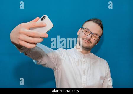 Un giovane uomo d'affari europeo, elegante, affaristico e sicuro, in occhiali e maglietta bianca, prende un selfie sul suo smartphone su un backgrou blu da parete Foto Stock