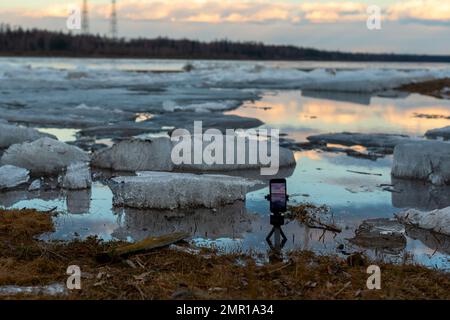 Lo schermo di un telefono cellulare spara un video della deriva di ghiaccio primaverile in piedi su un piccolo treppiede nel fiume, sullo sfondo di galleggianti di ghiaccio. Foto Stock