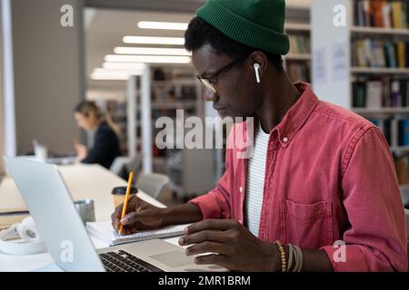 Un uomo afroamericano concentrato e senza emozioni che prende appunti nel notebook si siede al tavolo con il notebook Foto Stock
