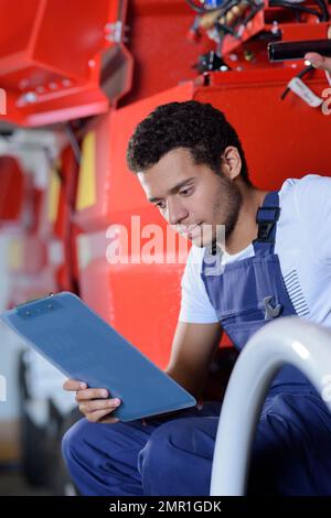 giovane meccanico maschio seduto sul veicolo passo lettura appunti Foto Stock