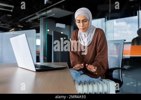 Gelare donna araba in ufficio, donna d'affari in hijab freddo riscaldamento interno e riscaldamento non funziona, utilizzando il notebook sul lavoro sul posto di lavoro, seduto cercando di riscaldare. Foto Stock