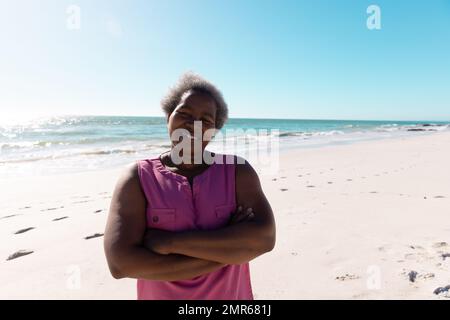 Donna anziana afroamericana con le braccia incrociate in piedi in spiaggia contro il mare e il cielo tranquilli Foto Stock