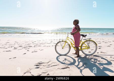 Vista laterale di una donna anziana afroamericana con bicicletta in piedi su una spiaggia sabbiosa contro il cielo limpido Foto Stock