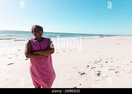 Donna anziana afroamericana con braccia incrociate in piedi su spiaggia sabbiosa sotto il cielo limpido nelle giornate di sole Foto Stock
