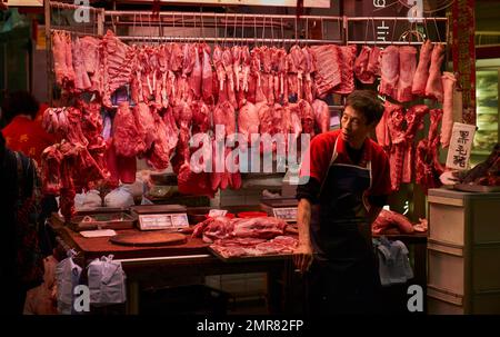 Un macellaio in un grembiule si trova di fronte alla carne cruda appesa al suo stand in un mercato all'aperto in una strada di Hong Kong. Foto Stock