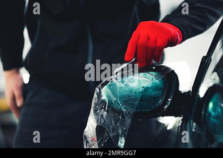 Specchio laterale rivestito con pellicola protettiva per verniciatura auto da un professionista in un guanto rosso. Procedura di dettaglio della vettura. Foto di alta qualità Foto Stock