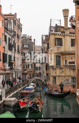 La scena dei canali di Venezia raffigura piccole barche e gondole in una zona residenziale di Venezia in inverno in una fresca giornata grigia con un popolo sulla strada Foto Stock