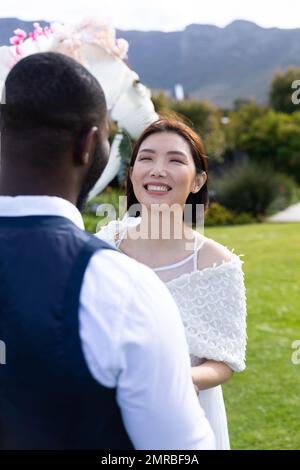 Verticale di felice sposa asiatica sorridente a sposo diverso a nozze all'aperto, copia spazio Foto Stock