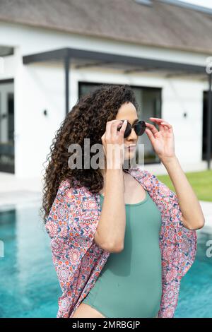 Verticale di donna biraciale in occhiali da sole e costume da bagno in piscina a casa vacanze, copia spazio Foto Stock