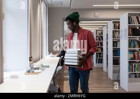 Studente africano che tiene un mucchio di libri e guarda lo schermo del computer portatile mentre studia in biblioteca Foto Stock
