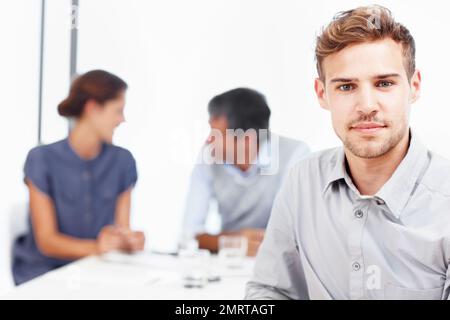 Il nostro team ottiene sempre il lavoro fatto. Primo piano ritratto di un giovane uomo d'affari fiducioso con i suoi colleghi seduti sullo sfondo. Foto Stock