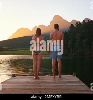 Ammirando una magnifica vista. Ripresa da dietro di una giovane coppia in costume da bagno in piedi su un molo al tramonto. Foto Stock