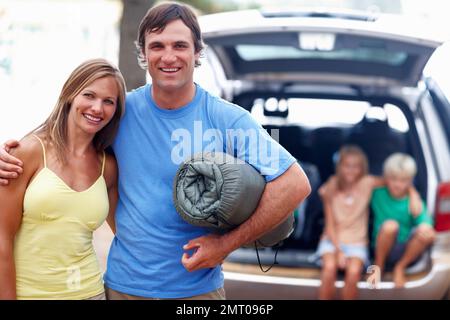 Coppia pronta per il campo. Ritratto di coppia con un uomo in possesso di materasso da campeggio e bambini seduti in auto. Foto Stock