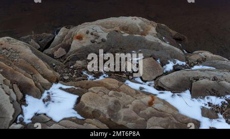 Foto di un affioramento roccioso sulla riva con acqua scura con neve e tracce di ruggine. Foto Stock