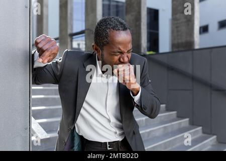 Attacco di tosse. Un uomo d'affari afro-americano in tuta sta tenendo sulla parete dell'ufficio fuori, tossendo duro, coprendo la bocca con la mano. sente dolore toracico. Foto Stock