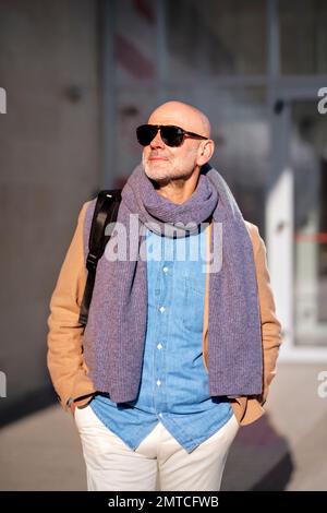 Uomo d'affari maturo in abiti casual camminando all'aperto da un edificio e godendosi una giornata di sole. Maschio caucasico che indossa occhiali da sole e scharf. Foto Stock