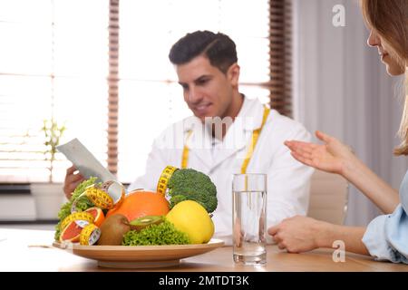 Nutrizionista consulenza paziente a tavola in clinica, concentrarsi su piastra con frutta, verdura e metro a nastro Foto Stock