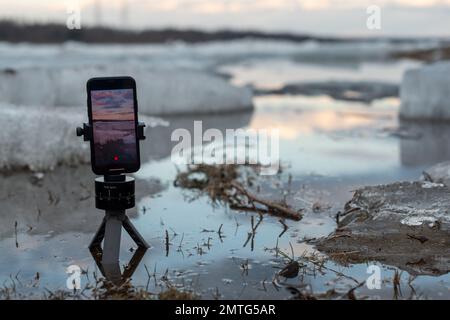 Un telefono cellulare scatta un video della deriva di ghiaccio primaverile in piedi su un piccolo treppiede in acqua, sullo sfondo di galleggianti di ghiaccio Foto Stock