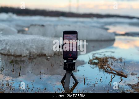 Un telefono cellulare scatta un video della deriva di ghiaccio primaverile in piedi su un piccolo treppiede in acqua, sullo sfondo di galleggianti di ghiaccio Foto Stock