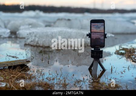 Lo schermo di un telefono cellulare spara un video della deriva di ghiaccio primaverile in piedi su un piccolo treppiede nel fiume, sullo sfondo di galleggianti di ghiaccio. Foto Stock