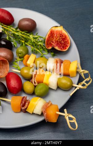 Formaggio, olive, jamon su spiedino, fichi e rametti di timo su piatto grigio. Vista dall'alto. Sfondo blu. Foto Stock