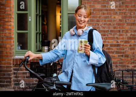 Giovane bella sorridente felice donna elegante in camicia blu che tiene e utilizza il suo telefono , mentre in piedi con la bicicletta vicino a muro di mattoni all'aperto Foto Stock