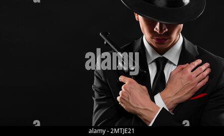 Elegante ritratto di gangster del 1940s con una pistola. Foto ravvicinata di un giovane uomo in tuta nera e cappello con una pistola su sfondo scuro. Foto Stock