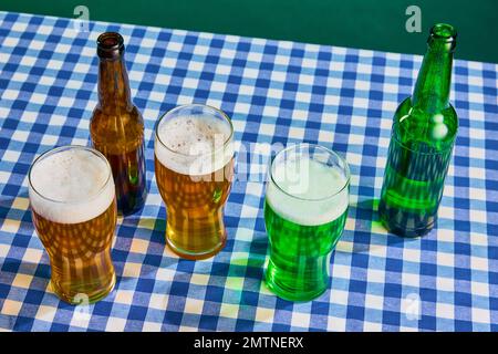 Vista dall'alto di bottiglie e bicchieri con birra magra e schiumosa verde su tovaglia a scacchi su sfondo verde. Concetto di celebrazione del giorno di san patrizio Foto Stock