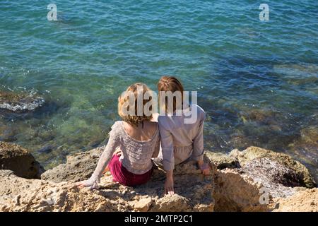 due ragazze adulte si siedono con le spalle sulle rocce sullo sfondo del mare turchese. Mamma e figlia adulta, fidanzate, sorelle in vacanza, Foto Stock
