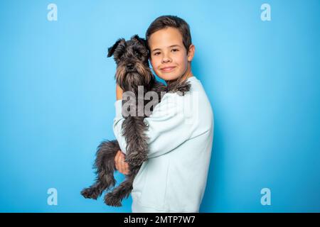 Il cane Schnauzer e il ragazzo sorridente la abbraccia con tenerezza e guardano la telecamera su sfondo giallo. Animali domestici. Emozioni della gente. Infanzia. Stile di vita. PET c Foto Stock