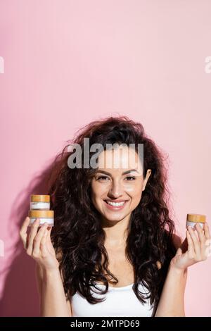 Curly giovane donna che tiene creme cosmetiche su sfondo rosa, immagine di scorta Foto Stock