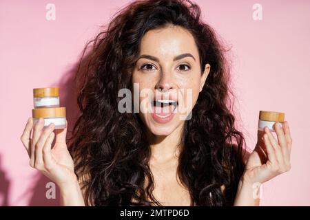 Donna curly eccitato tenere vasi con creme su sfondo rosa, immagine stock Foto Stock