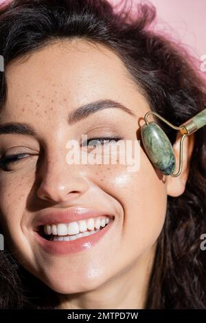 Vista ravvicinata di una donna sorridente incisa che utilizza un rullo di giada isolato su un'immagine rosa Foto Stock