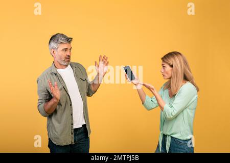 Concetto di truffa. Donna arrabbiata di mezza età che controlla il cellulare del marito, chiedendo spiegazioni, l'uomo che sparge le mani Foto Stock